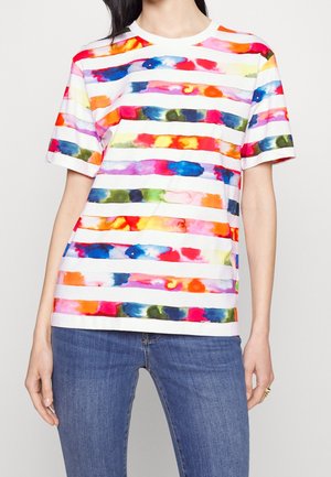 Kobieta ubrana w białą krótką koszulkę z poziomymi paskami w stylu tie-dye w kolorach czerwonym, niebieskim, żółtym, różowym, zielonym i pomarańczowym, w połączeniu z niebieskimi dżinsami.