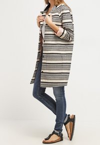 Cappotto a righe con motivi orizzontali in bianco e nero, abbinato a jeans blu e sandali neri con suole testurizzate.