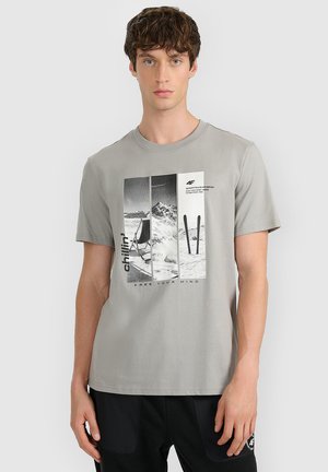 Młody mężczyzna w jasnoszarej koszulce z grafiką góry i nart oraz słowami "chillin' free your mind."
