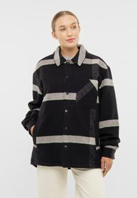 Černá a šedá pruhovaná bunda s měkkou texturou, zapínání na knoflíky a dlouhými rukávy. Obsahuje náprsní kapsu a velký límec.