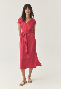 Donna in piedi con un vestito midi rosso a maniche corte, una cintura in vita e sandali beige con punta aperta e piatti, su uno sfondo neutro.