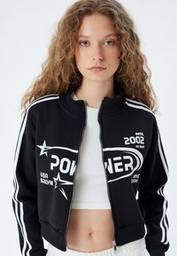Giacca nera con zip, con strisce bianche sulle maniche, presenta testo grafico e un design a stella. Realizzata in tessuto morbido, taglio fitted, polsini a coste.