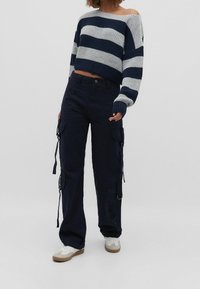 Laivastonsiniset cargo-housut sivutaskuilla ja säädettävillä kiristysnyöreillä; yhdistetty harmaan ja laivastonsinisen raidallisen lyhyen neulepaidan kanssa. Valkoiset tennarit.