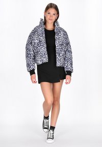 Felső részen rövidített, világosszürke színű pufi dzseki fekete leopárdmintával. Magas gallérral, cipzáras záródással és bordázott mandzsettákkal rendelkezik.