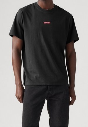Mand iført en sort Levi’s T-shirt med korte ærmer og sorte jeans, stående mod en ensfarvet lys baggrund.