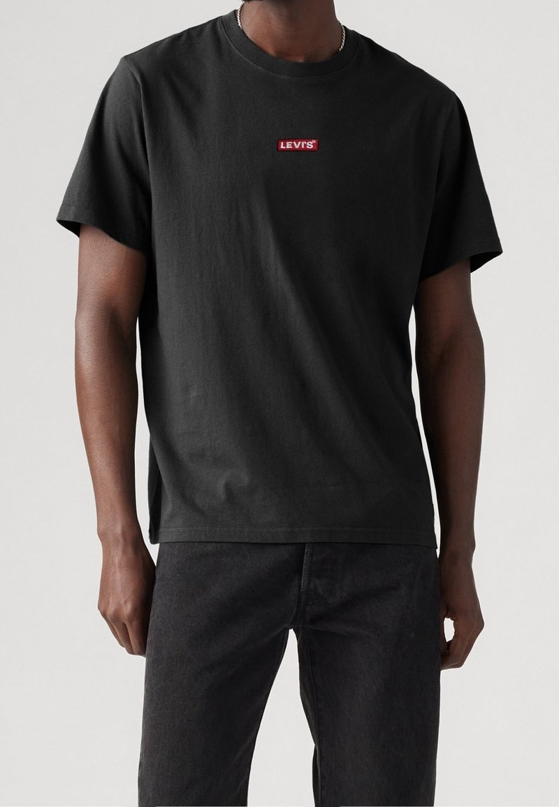 Homme portant un T-shirt noir Levi’s à manches courtes et un jean noir, debout devant un fond clair uni.