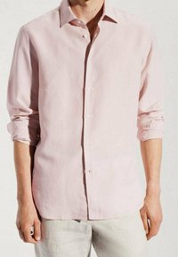 Camisa de lino rosa claro con cuello abotonado, mangas largas remangadas, corte recto y textura suave. Botones blancos acentúan la parte delantera.