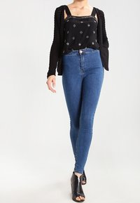 Black sleeveless top with patterned accents, paired with high-waisted blue jeans. Model wears black open-toe high-heeled shoes.
