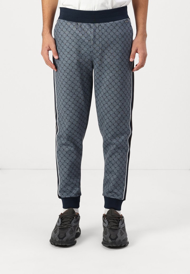Blaue, gemusterte Sweatpants mit geometrischem Design, elastischem Bund und seitlichen Streifen. Abgeschlossen mit eng anliegenden Bündchen und kombiniert mit dunklen Schuhen.