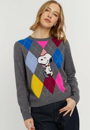 Vrouw draagt een grijze argyle trui met kleurrijke ruiten en het personage Snoopy in het midden, gecombineerd met donkerblauwe spijkerbroek.