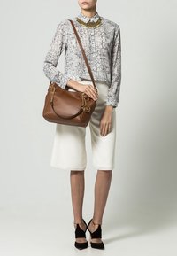 Brązowa skórzana torba na ramię z złotym okuciem, okrągłym logo i teksturowanym wzorem. Noszona z wzorzystą koszulą i białymi kulotami.