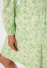 Green floral patterned dress with a light background, featuring long sleeves and a button front closure. The fabric is textured and lightweight.