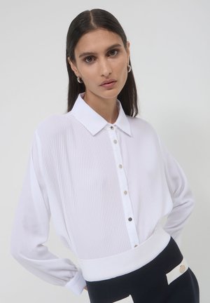 Donna con lunghi capelli castani che indossa orecchini a cerchio d'argento, una blusa bianca a pieghe con bottoni e una gonna in bianco e nero, in piedi davanti a uno sfondo bianco.