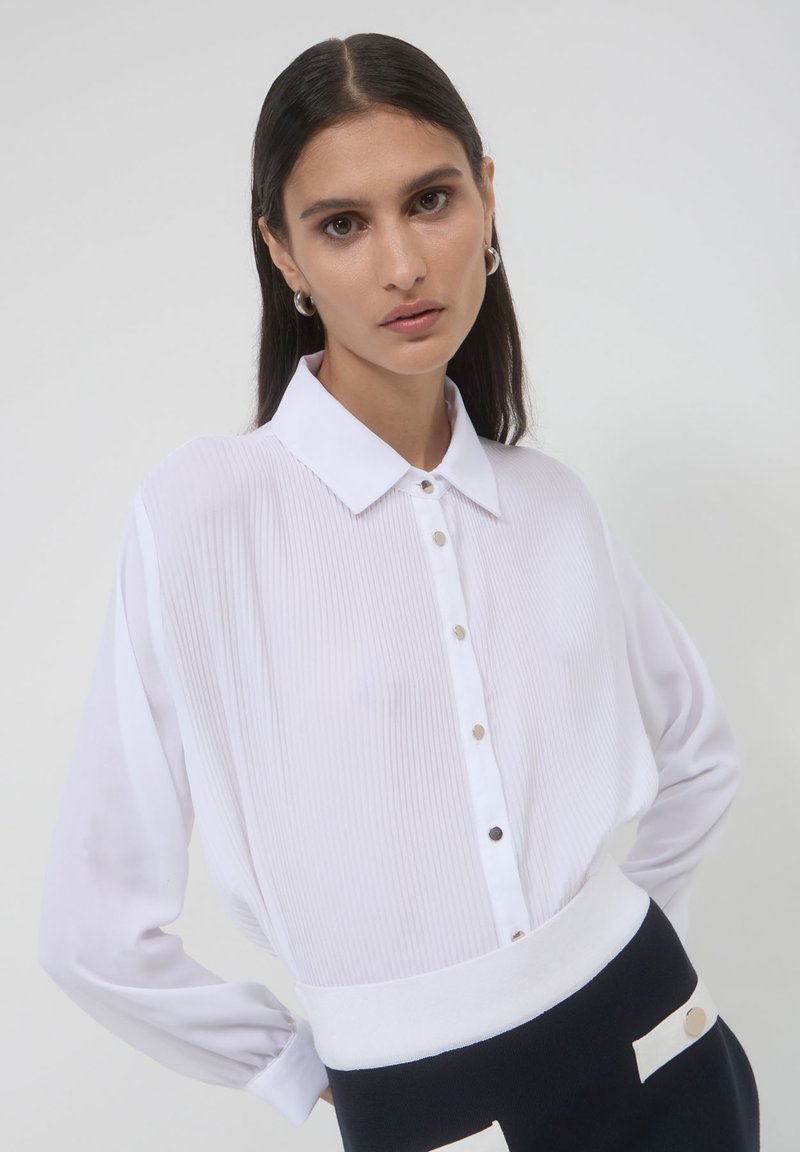 Donna con lunghi capelli castani che indossa orecchini a cerchio d'argento, una blusa bianca a pieghe con bottoni e una gonna in bianco e nero, in piedi davanti a uno sfondo bianco.