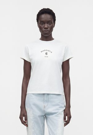 Femme aux cheveux courts portant un t-shirt blanc avec le logo Margiela 6 et un jean en denim bleu clair, debout devant un fond blanc uni.