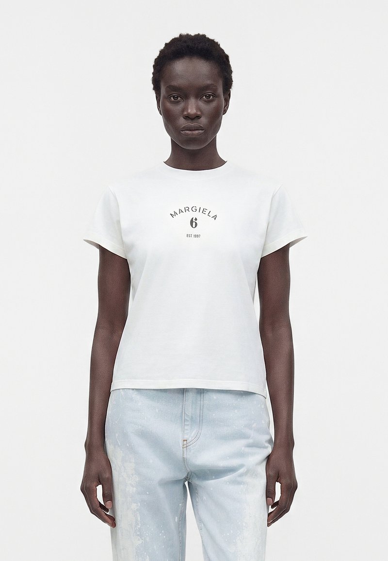 Γυναίκα με κοντά μαλλιά που φοράει λευκό t-shirt με το λογότυπο Margiela 6 και ανοιχτό μπλε τζιν, στέκεται σε λευκό φόντο.