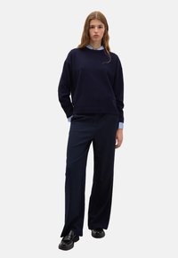 Maglione blu navy profondo con collo rotondo, vestibilità rilassata e maniche lunghe, indossato sopra una camicia collare azzurra. Abbinato a pantaloni navy a gamba larga.