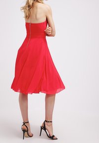 Rød, stropløs kjole lavet af let, flydende stof. Knælængde med en tætsiddende overdel og udvidet skirt. Har en lynlås i ryggen. Sorte stiletto hæle.