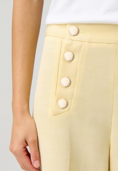 Un pantalon jaune en tissu texturé présente quatre grands boutons métalliques sur le côté. Une main repose près d'une poche latérale.