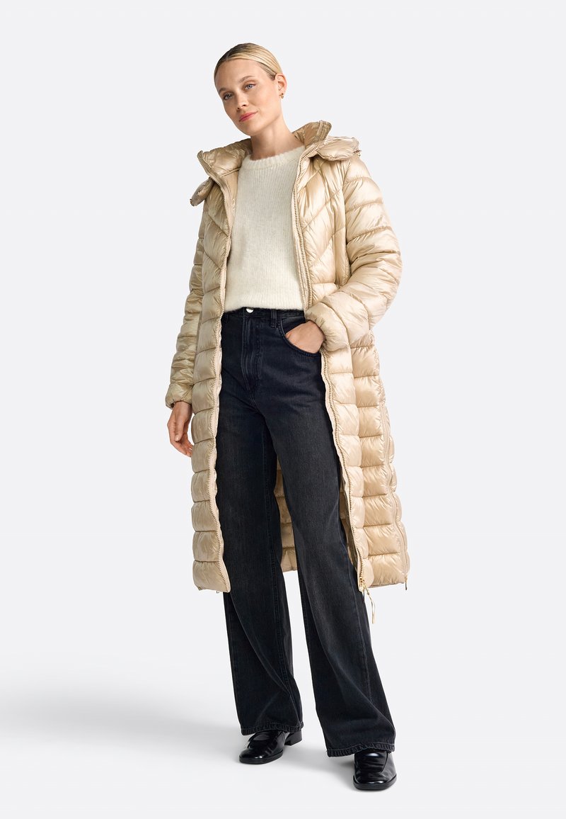 Lang, lichtgewicht, beige puffercoat met horizontaal gestikte ontwerpen, ritssluiting en capuchon, gedragen over een crèmekleurige trui en zwarte spijkerbroek.