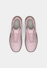 Sapatilhas atléticas cor-de-rosa com textura de camurça, apresentando uma língua em malha cinza, uma marcação dourada e uma sola de borracha castanha. Cadarços na mesma cor.