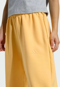 Osoba oblečená v svetločervených teplákoch s elastickým pásom a embosovaným logom Adidas, doplnená o šedý krátky top, stojí.