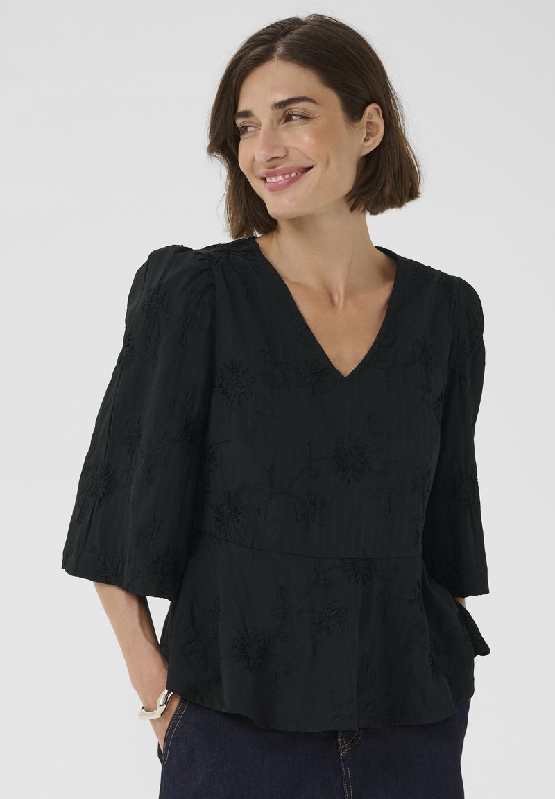 Schwarze Peplum-Bluse mit V-Ausschnitt, strukturiertem Stoff, floraler Stickerei und Dreiviertelärmeln, kombiniert mit dunklem Denim.