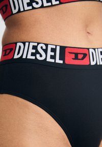 Culotte noire avec une large ceinture comportant un logo "DIESEL" répété en blanc et rouge. Texture de tissu lisse avec des détails de couture minimaux.