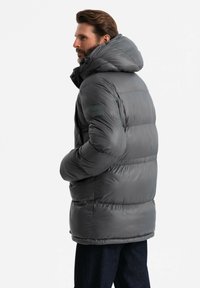 Giacca puffer di colore grigio scuro con cappuccio, texture trapuntata e tasche laterali. Presenta una piccola etichetta con logo sul braccio superiore.