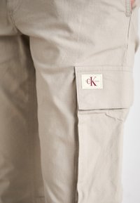 Pantalones cargo beige hechos de una tela texturizada, con un bolsillo lateral y una etiqueta de logo en rojo y crema cerca de la cintura.