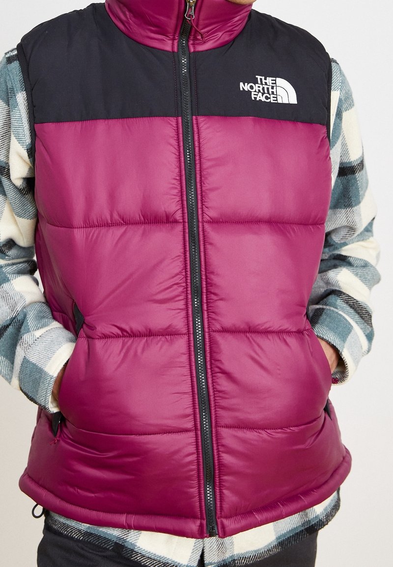 Personne portant une veste matelassée North Face bordeaux et noire sur une chemise à carreaux verte et blanche, les mains dans les poches de la veste.