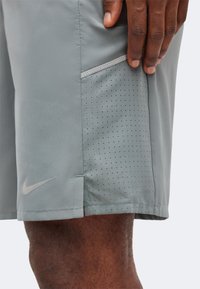 Κοντινό πλάνο ενός χεριού που ακουμπάει σε γκρι αθλητικό σορτς με οπές για αερισμό και το λογότυπο της Nike κοντά στην άκρη.