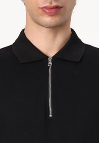 Tricou polo negru cu guler și detaliu cu fermoar la jumătate, având un material texturat și un design elegant, minimalist.
