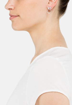 Silberne Creolen mit einer zarten Schleife und kleinen Kunstperlen, vor einem hellen Hintergrund, getragen von einer Person in einem weißen Hemd.