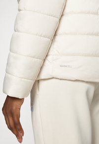 Light beige puffer jacket with quilted sections and a smooth texture. "WARMCELL" logo is visible near the hem, paired with beige sweatpants.