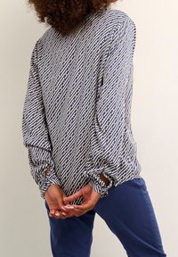 Langærmet bluse i marineblåt og creme med geometrisk mønster; løs pasform med rynkede ærmer og udskæringsdetaljer ved håndleddene. Kombineret med marineblå bukser.