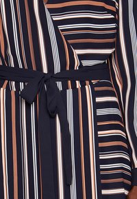 Sukienka kopertowa w granatowym kolorze z pionowymi paskami w pomarańczowym i białym kolorze. Posiada wiązanie w talii oraz długie rękawy, wykonana z gładkiego materiału.