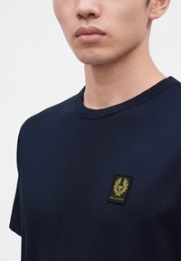 Νεαρό άτομο που φοράει μπλε ναυτικό T-shirt με ένα μικρό πατς λογότυπο Belstaff στο αριστερό στήθος, πάνω σε ένα απλό φωτεινό φόντο.