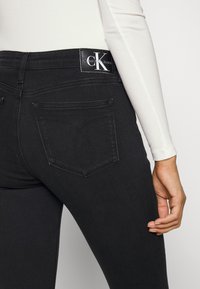 Jeans de mezclilla negra con un diseño ajustado, que cuentan con bolsillos traseros y una etiqueta con el logotipo de Calvin Klein en la cintura. Textura suave.