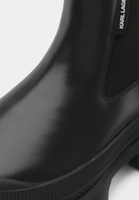 Bota de tornozelo de borracha preta com acabamento brilhante, painéis laterais elásticos e sola texturizada com detalhe de marca ao longo da borda.