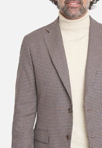 Blazer a cuadros marrones con solapa con muesca, dos botones y bolsillos laterales, usado sobre un suéter de cuello alto de color beige claro.