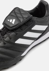 Scarpa da calcio nera in pelle sintetica, con linguetta trapuntata, tre strisce bianche e tacchetti neri in gomma sulla suola.