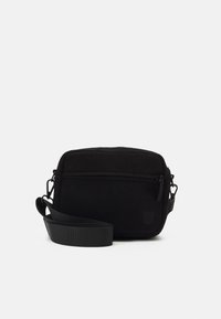 Black corduroy crossbody bag with a single zipper compartment, adjustable strap, and a textured surface. Features a subtle logo.