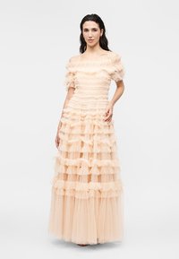 Naine, kes kannab põrandapikkust õlgadeta virsikuvärvi kleiti, millel on kihilised tüllvolangid, seisab ühevärvilise valge tausta ees.