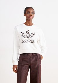 Hvit cropped sweatshirt med leopardmønstret Adidas-logo, kombinert med brune mønstrede bukser, med elastisk midje og sidestriper.