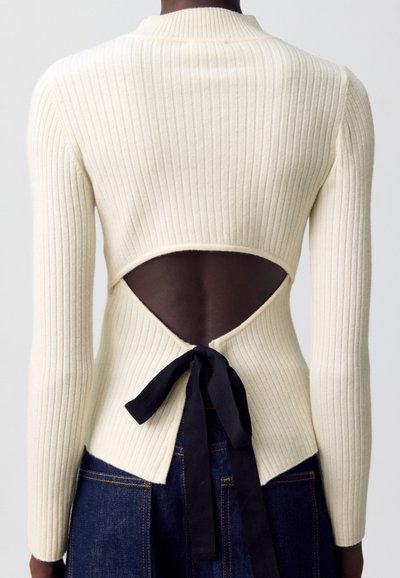 Vue arrière d'une personne portant un haut à manches longues en crème à côtes avec un dos ouvert noué par un ruban noir, associé à un jean en denim bleu foncé.