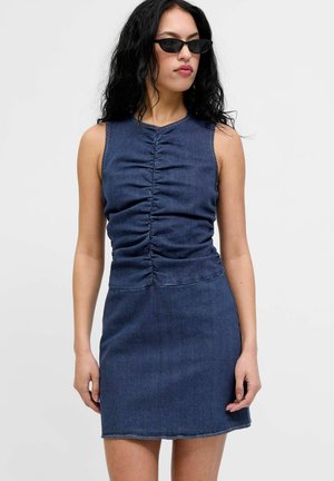 Femme aux longs cheveux noirs portant des lunettes de soleil noires et une robe en denim sombre sans manches, froncée, sur un fond uni.