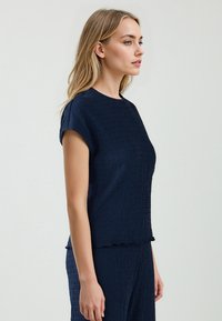 Navy-blaues, strukturiertes Oberteil mit Rundhalsausschnitt und kurzen Ärmeln, das ein wellenförmiges Muster und einen Rüschensaum aufweist. Glatte Stoffoberfläche.