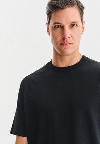 Mężczyzna z krótkimi brązowymi włosami i zielonymi oczami, ubrany w prostą czarną koszulkę typu crew-neck na białym tle.