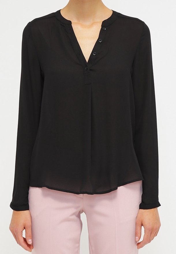 Blouse noire à manches longues avec col en V boutonné portée avec un pantalon rose clair, vue du cou aux hanches sur une personne debout.